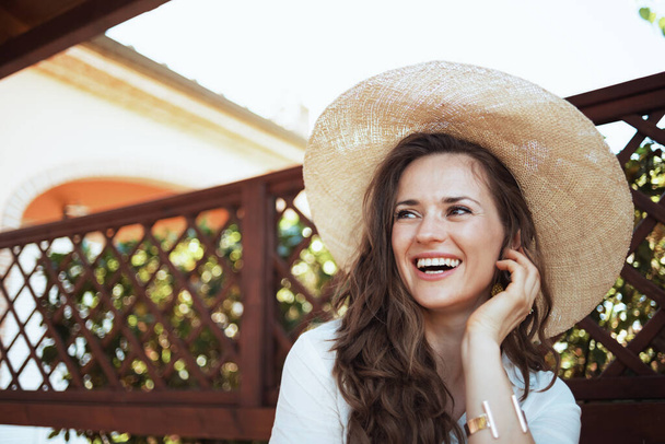 щаслива модна домогосподарка середнього віку в білій сорочці з капелюхом на терасі готельного будинку
. - Фото, зображення