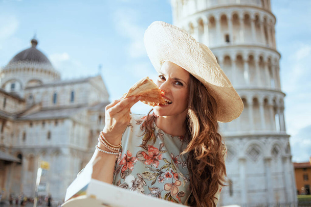smiling young solo traveller woman in floral dress with pizza and hat near Leaning Tower in Pisa, Italy. - Foto, imagen