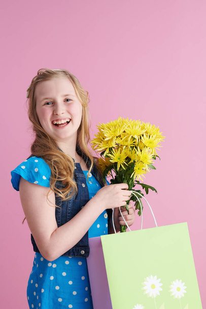 happy stylish child with long wavy blond hair with yellow chrysanthemums flowers and green shopping bag isolated on pink. - Foto, Bild