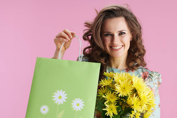Porträt einer lächelnden trendigen Frau mit langen welligen brünetten Haaren mit gelben Chrysanthemen-Blumen und grüner Einkaufstasche auf rosa Hintergrund. - Foto, Bild