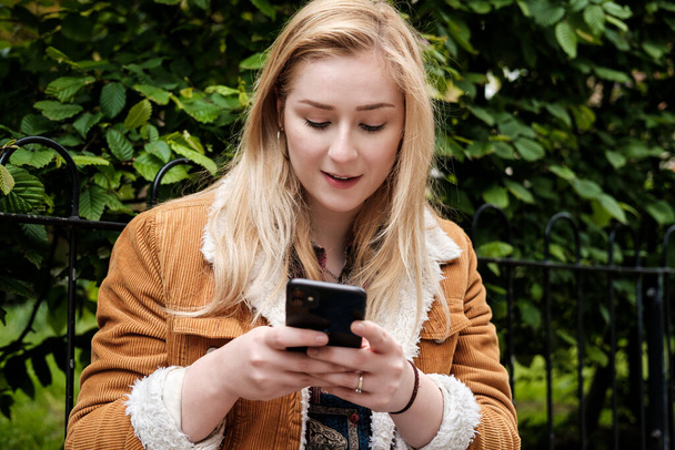 Πορτρέτο μιας νεαρής γυναίκας που χρησιμοποιεί το smartphone της. Φοράει καφέ παλτό και χτυπάει την οθόνη... - Φωτογραφία, εικόνα