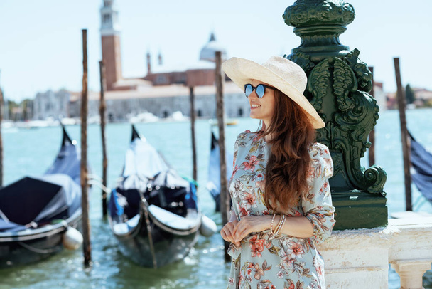 sonriente mujer viajera solista de moda en vestido floral con gafas de sol y sombrero teniendo recorrido a pie en terraplén en Venecia, Italia. - Foto, imagen