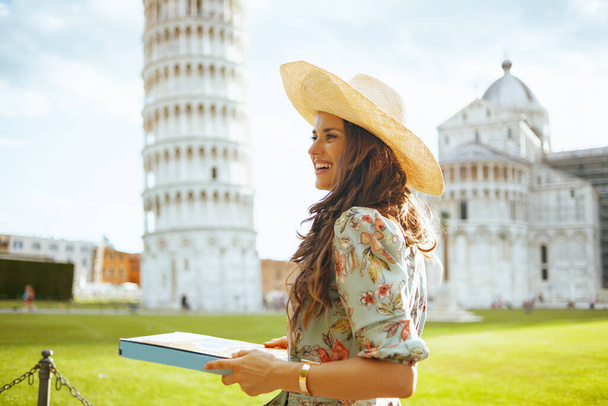 happy elegant middle aged traveller woman in floral dress with pizza and hat near Leaning Tower in Pisa, Italy. - Fotó, kép