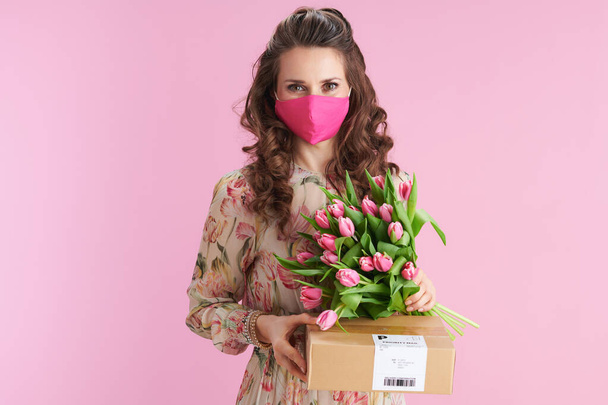 young woman with long wavy brunette hair with tulips bouquet, pink mask and parcel against pink background. - Фото, зображення