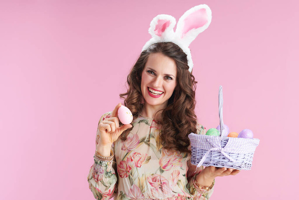 middle aged woman with long wavy brunette hair with bunny ears and basket of easter eggs isolated on pink background. - Foto, Imagen