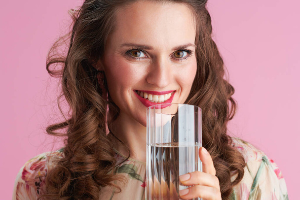 Portrait of happy trendy female with long wavy brunette hair with glass of water against pink background. - Fotoğraf, Görsel