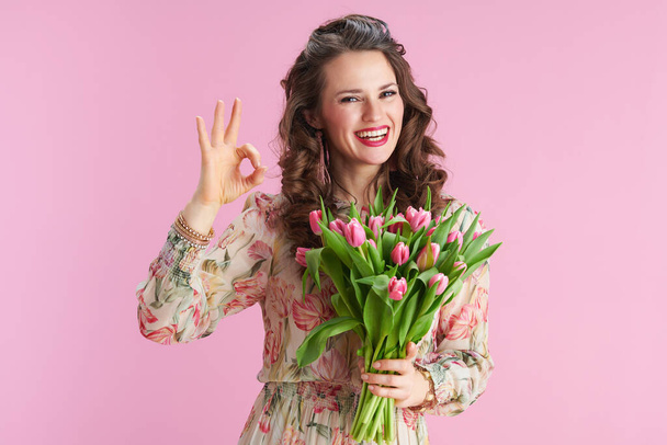 happy elegant middle aged woman in floral dress with tulips bouquet showing ok gesture isolated on pink. - Photo, Image