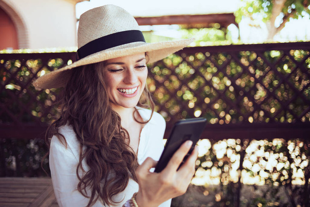 happy stylish 40 years old woman in white shirt with hat having virtual meeting on a smartphone in the terrace. - 写真・画像