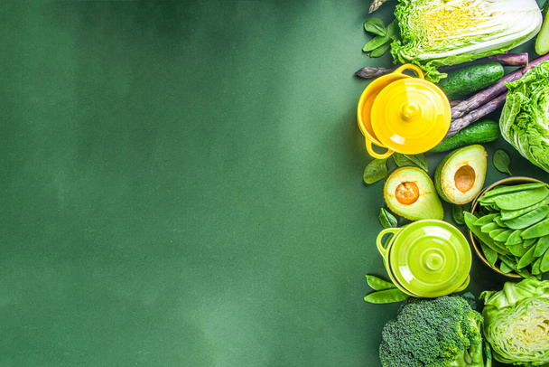 Various organic green spring vegetable ingredients with empty colorful portioned cooking pots on dark green kitchen table, top view. Diet healthy soup, stew food preparation concept - 写真・画像