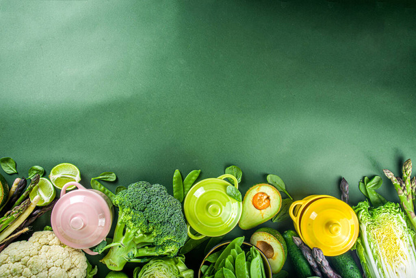 Verschiedene organische grüne Frühlingsgemüsezutaten mit leeren, bunt portionierten Kochtöpfen auf dunkelgrünem Küchentisch, Draufsicht. Diät gesunde Suppe, Eintopf Zubereitungskonzept - Foto, Bild