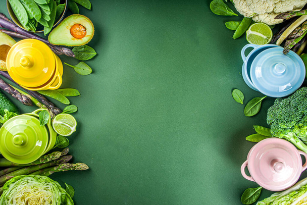 Разнообразная органическая зеленая весна, ингредиенты с пустыми цветными порционными кухонными горшками на темно-зеленом кухонном столе, вид сверху. Диетический здоровый суп, концепция приготовления рагу - Фото, изображение