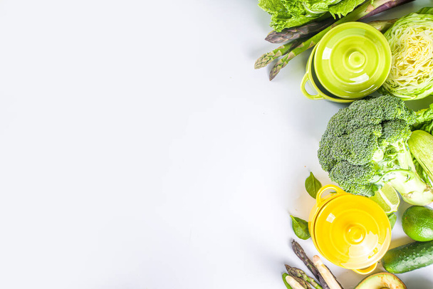 Διάφορα βιολογικά πράσινα ανοιξιάτικα φυτικά συστατικά με άδεια πολύχρωμα μαγειρικά σκεύη σε λευκό τραπέζι κουζίνας, θέα στην κορυφή. Δίαιτα υγιεινή σούπα, στιφάδο - Φωτογραφία, εικόνα
