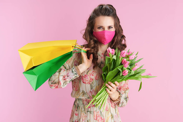 stylish female in floral dress with tulips bouquet, pink mask and shopping bags isolated on pink. - Фото, зображення