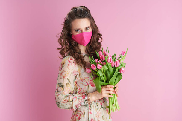 joven mujer de 40 años con el pelo largo y ondulado morena con tulipanes ramo y máscara rosa aislado en rosa. - Foto, imagen
