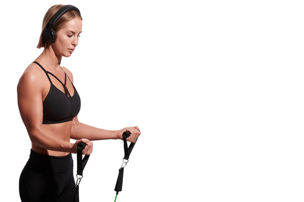 Sterke gespierde sportvrouw in hoofdtelefoon die traint met een weerstandsband over een witte geïsoleerde achtergrond - Foto, afbeelding