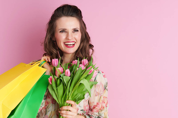 happy stylish female with long wavy brunette hair with tulips bouquet and shopping bags isolated on pink. - Φωτογραφία, εικόνα