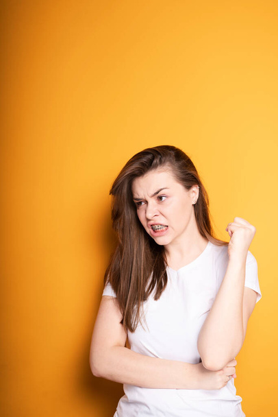 Annoyed young girl in a white t-shirt with braces suffers from pain in the teeth while standing on a yellow background - Fotó, kép