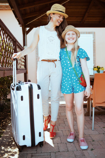 Full length portrait of smiling stylish mother and daughter tourists in hats with wheel bags and keys in the terrace of guest house hotel. - Zdjęcie, obraz