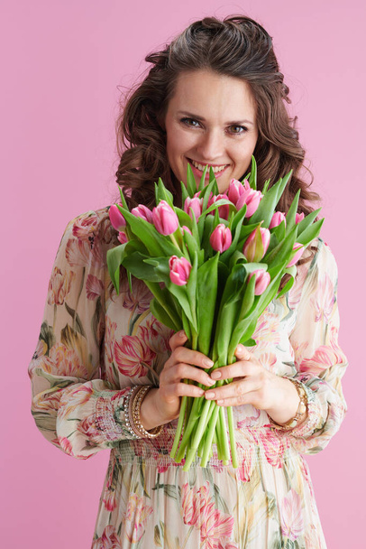 smiling young female in floral dress with tulips bouquet isolated on pink background. - Фото, изображение