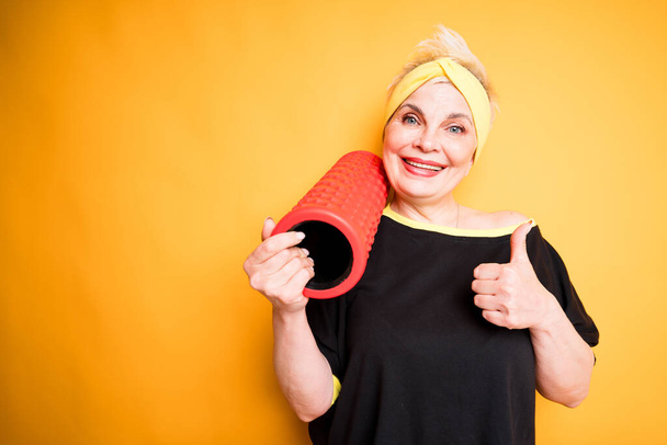 Cheerful elderly woman in sportswear holds fascia in hand while lifting thumb up - Fotografie, Obrázek