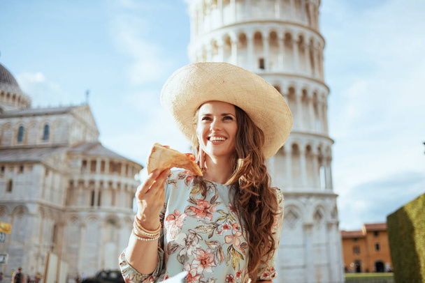 sonriente mujer viajera con estilo en vestido floral con pizza y sombrero en piazza dei miracoli en Pisa, Italia. - Foto, imagen