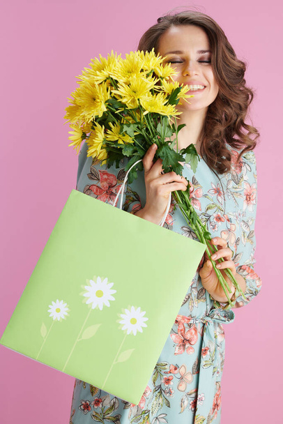 happy young woman with long wavy brunette hair with yellow chrysanthemums flowers and green shopping bag isolated on pink background. - Fotó, kép