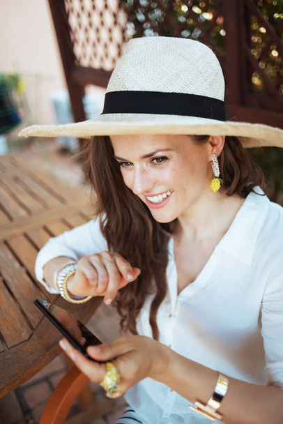happy trendy housewife in white shirt with hat sitting at the table sending text message using smartphone in the patio. - Фото, зображення