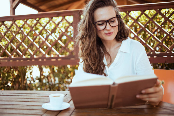 elegant woman in white shirt with cup of coffee and eyeglasses sitting at the table reading book in the patio of guest house hotel. - Фото, изображение