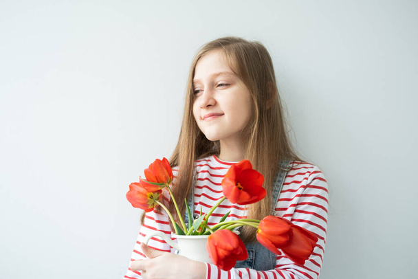 heureuse adolescente avec les cheveux longs tenant vase avec des tulipes rouges debout contre le mur blanc. - Photo, image