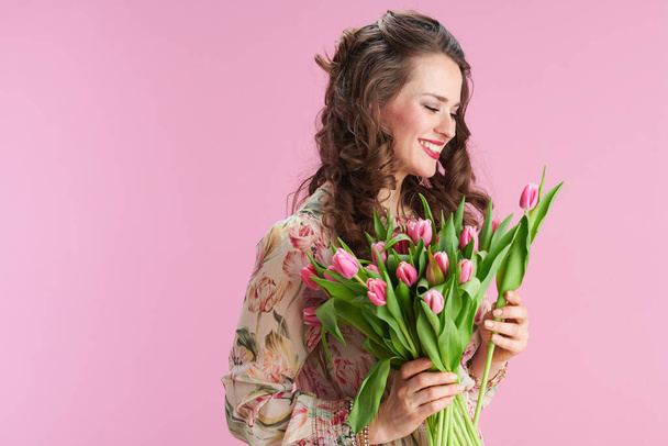 smiling trendy middle aged woman in floral dress with tulips bouquet isolated on pink background. - Fotó, kép