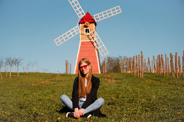 Beautiful young woman sits on the grass against the backdrop of the mill - Photo, Image