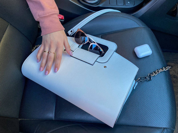 Witte vrouwelijke mooie modieuze leren tas en zonnebril met draadloze hoofdtelefoon en vrouwelijke hand in de auto. - Foto, afbeelding
