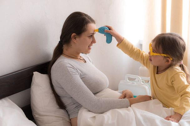 little girl wearing uniform playing doctor, nurse with young mum, nanny in bedroom, checking mother throat, measures temperature, take care, family spend leisure time at home together role play game - Foto, immagini