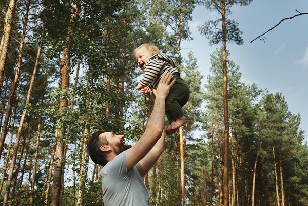 Happy father holds his baby high in the air against the backdrop of forest and tall pines. - Photo, Image