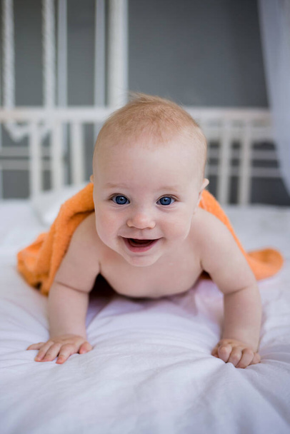 um menino feliz jaz em uma toalha de laranja na cama no quarto e olha para a câmera. Orientação vertical - Foto, Imagem