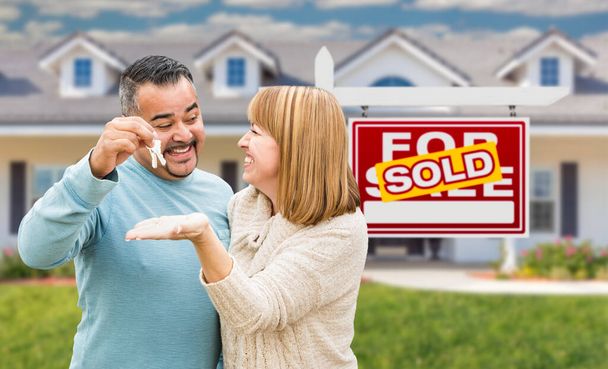 Gemischtes Rassenpaar vor verkauftem Immobilienschild und Haus mit Schlüsseln. - Foto, Bild