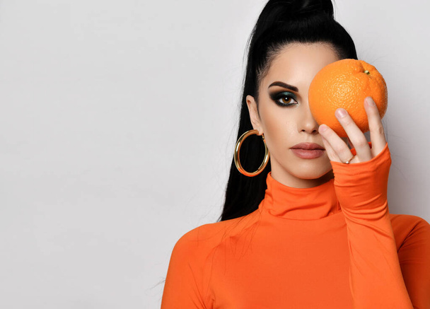 Young brunette woman in modern orange turtleneck and earrings holds an orange in front of her face over light background - Foto, imagen