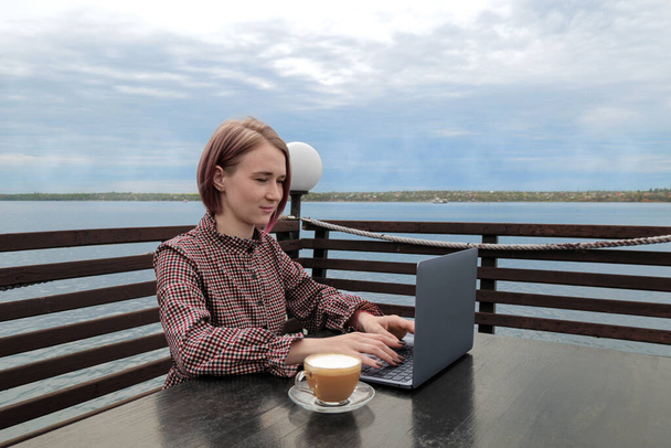 Una ragazza lavora per un portatile per strada. seduto a tavola a bere caffè con una splendida vista sul fiume. - Foto, immagini