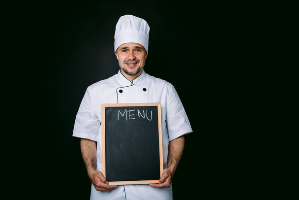 Chef cuisinier en veste et chapeau de cuisine souriant, tenant une piazarra avec le menu, sur fond noir - Photo, image