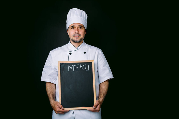 Chef cuisinier avec veste et chapeau de cuisine, tenant une piazarra avec le menu, sur fond noir - Photo, image