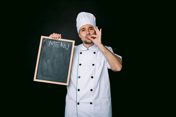 Chef cuisinier vêtu d'une veste et d'un chapeau de cuisine, tient une piazarra avec le menu et fait un bon geste alimentaire, sur fond noir - Photo, image