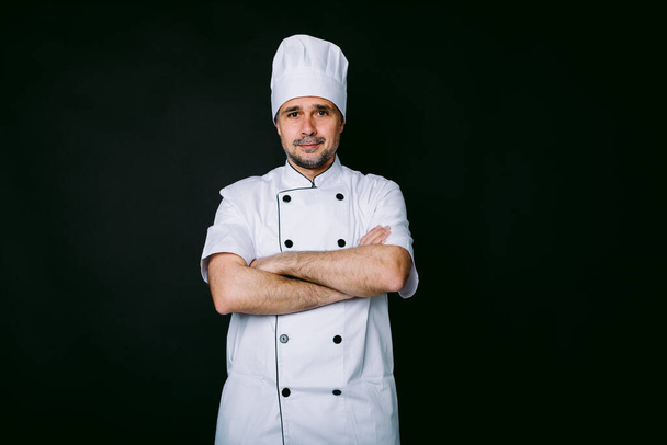Cocinero con chaqueta y sombrero de cocina, posa con los brazos cruzados sobre fondo negro - Foto, Imagen