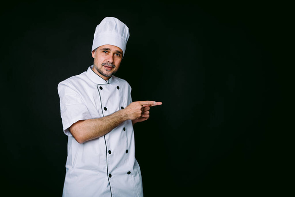 Szakács szakács visel kabát és konyhai kalap mutató jobbra fekete háttér fekete háttér - Fotó, kép