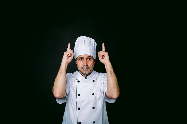 Μάγειρας σεφ φορώντας σακάκι και καπέλο κουζίνας που δείχνει προς τα πάνω σε μαύρο φόντο - Φωτογραφία, εικόνα