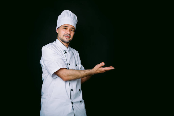 Chef cocinero vistiendo chaqueta y sombrero de cocina apuntando a la derecha sobre fondo negro - Foto, Imagen