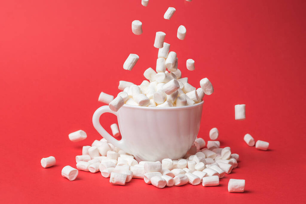 A large number of small marshmallows fall into a white cup. A sweet treat. - Фото, изображение