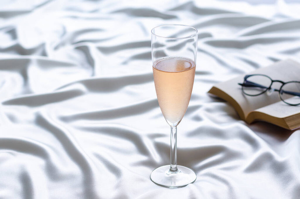 Een glas Rose wijn op golvende satijnen doek met boek en bril. Blijf thuis concept. - Foto, afbeelding