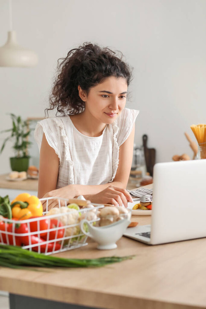 Young woman using laptop while eating in kitchen - Fotoğraf, Görsel