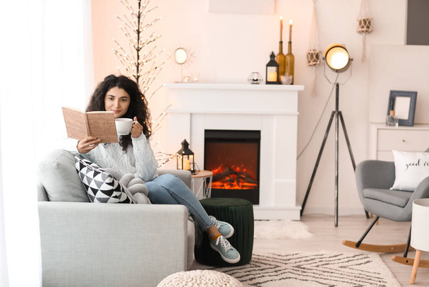 Beautiful woman drinking tea and reading book near fireplace at home - Photo, Image