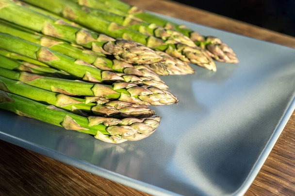 Close-up detail bunch of fresh raw green asparagus plant harvested at season on blue cerami plate kitchen background. Healthy vegeterain gourmet vegetable food. Health vegan diet nutrition concept - Photo, Image